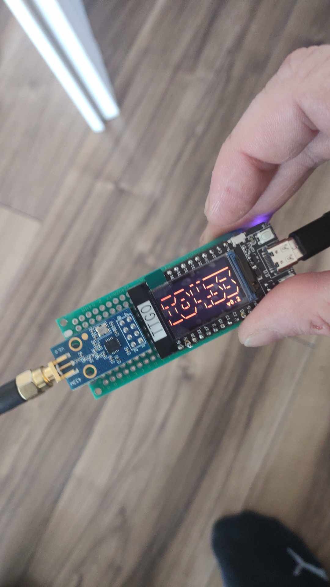 Zielona płytka drukowana z wyświetlaczem LED wskazującym cyfry, podłączona do kabla USB i anteny, trzymana w dłoni na tle drewnianej podłogi.