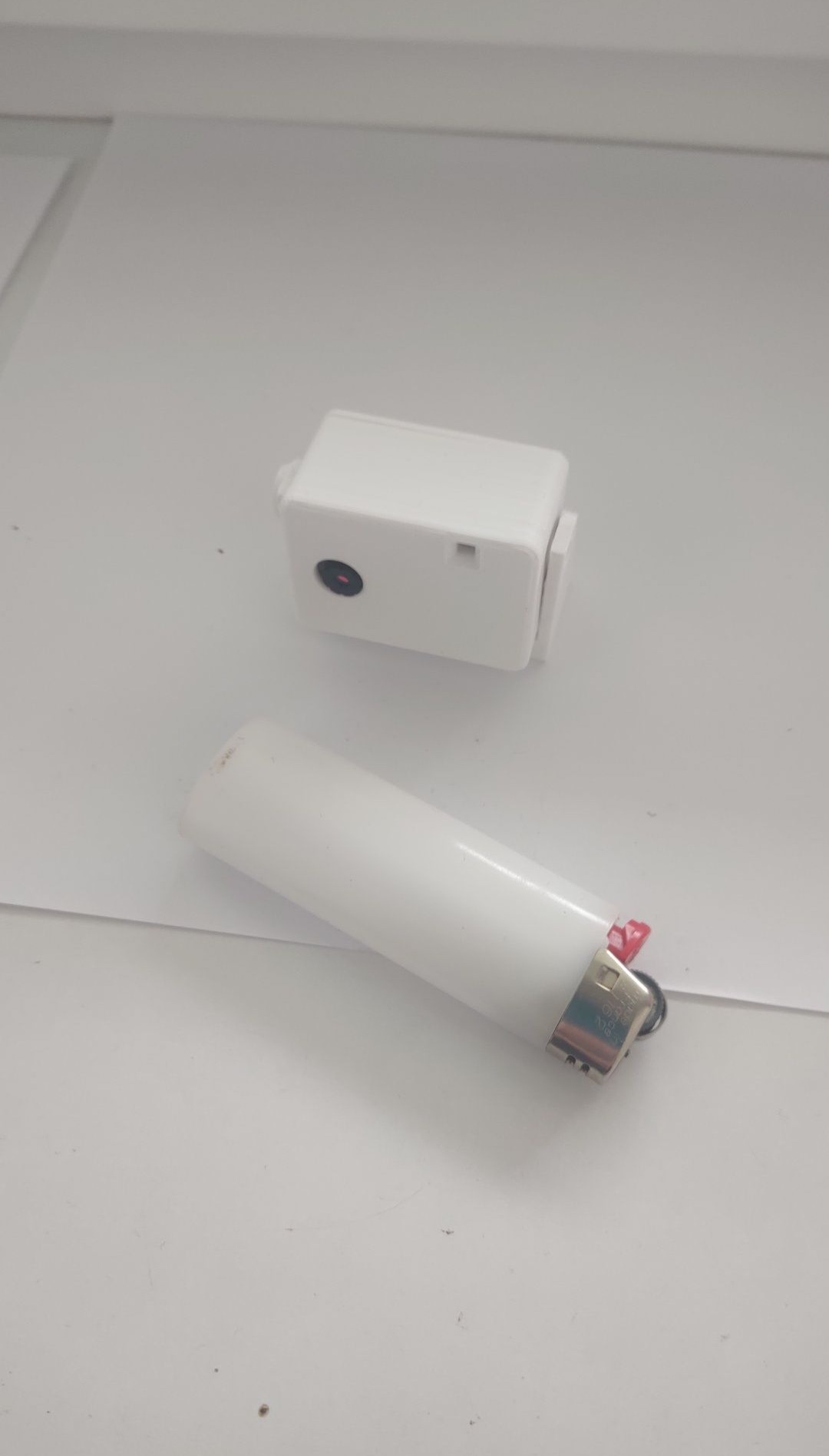 Mała, biała kamera monitoringu obok białej zapalniczki na białym tle, ukazująca kompaktowe rozmiary urządzenia.