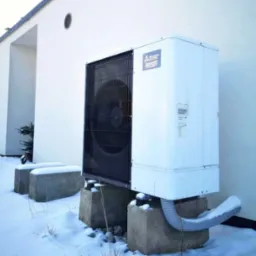 Zewnętrzna jednostka pompy ciepła Mitsubishi Zubadan, zamontowana na betonowych blokach w zimowym krajobrazie, z widoczną rurą odprowadzającą skropliny pokrytą śniegiem.