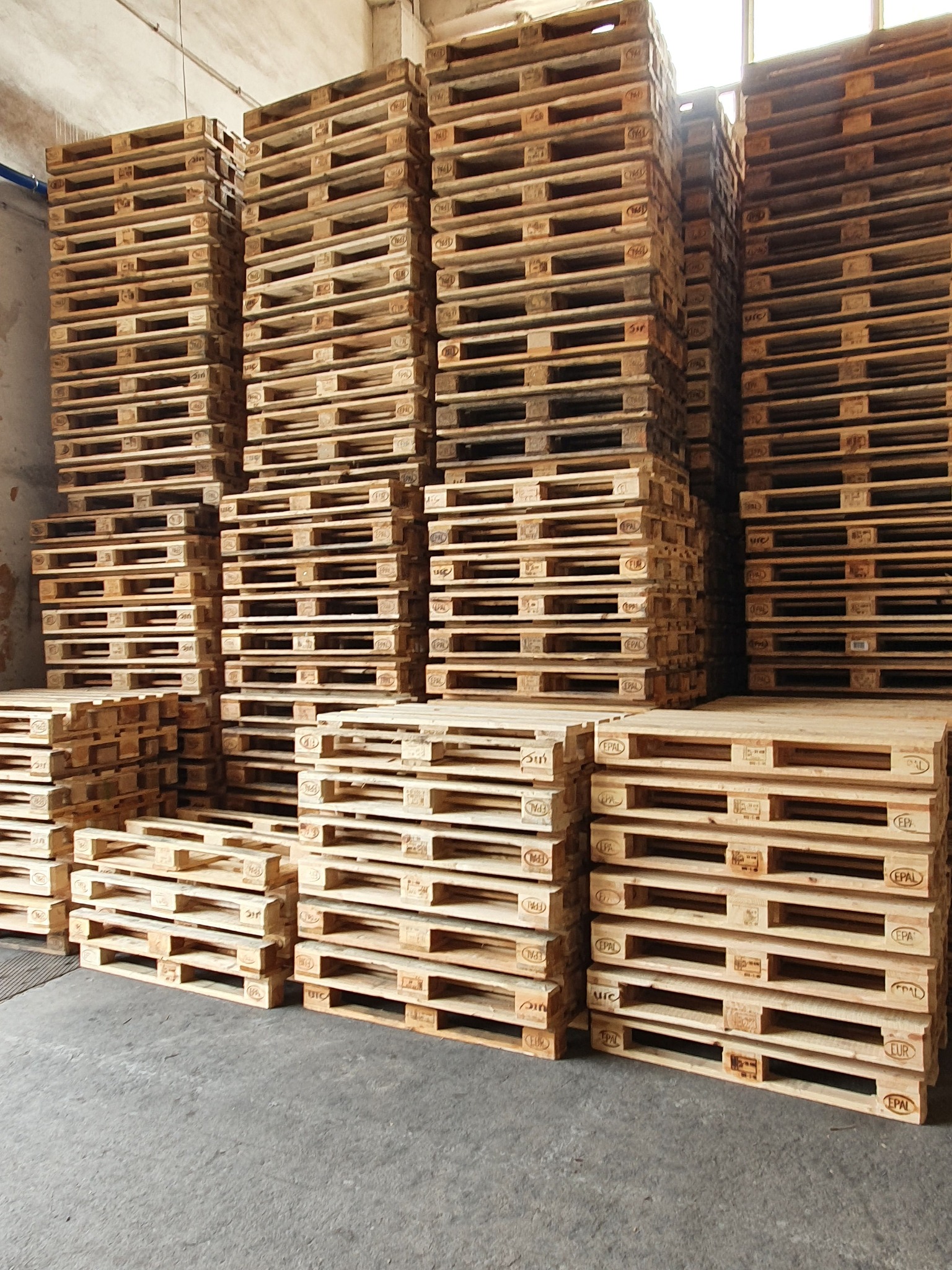 Stosy jasnych, drewnianych palet EPAL ułożone jeden na drugim w magazynie, widoczne oznaczenia na paletach.