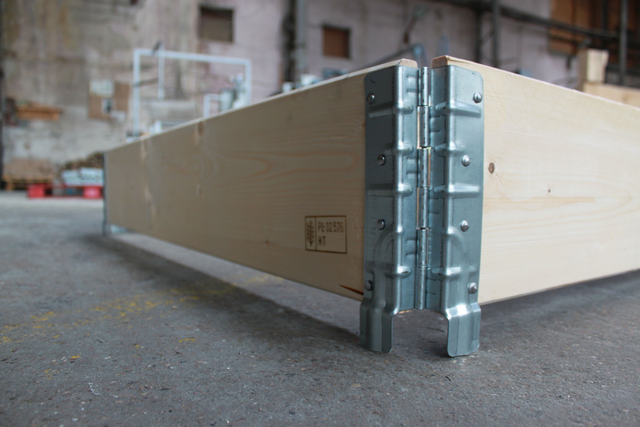 Fragment drewnianej ramy połączonej metalowymi kątownikami, widoczny znak certyfikacji na drewnie, surowy beton na podłożu, magazyn w tle.