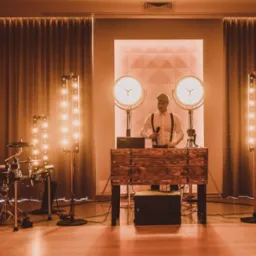 Wokalista w kaszkiecie i szelkach za drewnianym pulpitem DJ-a, mikrofon w dłoni, otoczony oświetleniem scenicznym i perkusją, ciepłe światło.