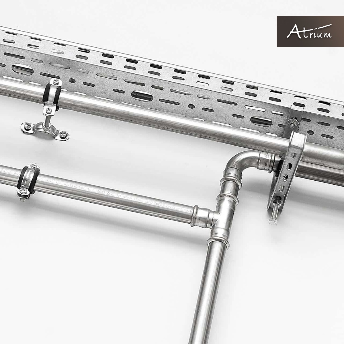 Metalowe rury wentylacyjne z mocowaniami na tle białej ściany, widoczne logo firmy Atrium w prawym górnym rogu.