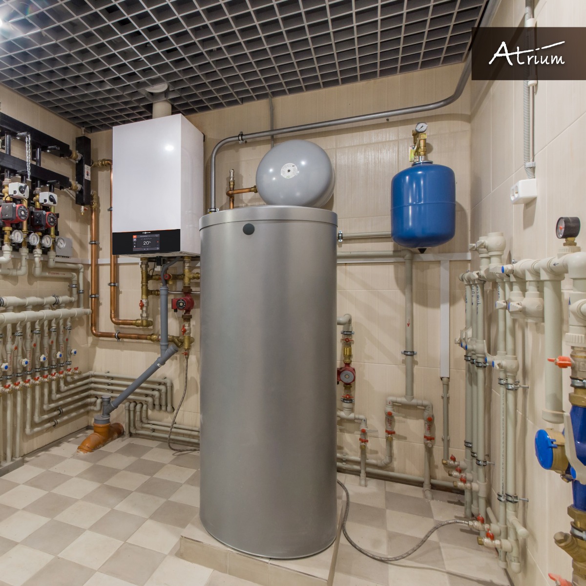 Kotłownia z nowoczesnym kotłem gazowym, zasobnikiem ciepłej wody i rozbudowaną instalacją hydrauliczną w pomieszczeniu wyłożonym kafelkami z kratownicą sufitową, widoczny firmowy logotyp w prawym...