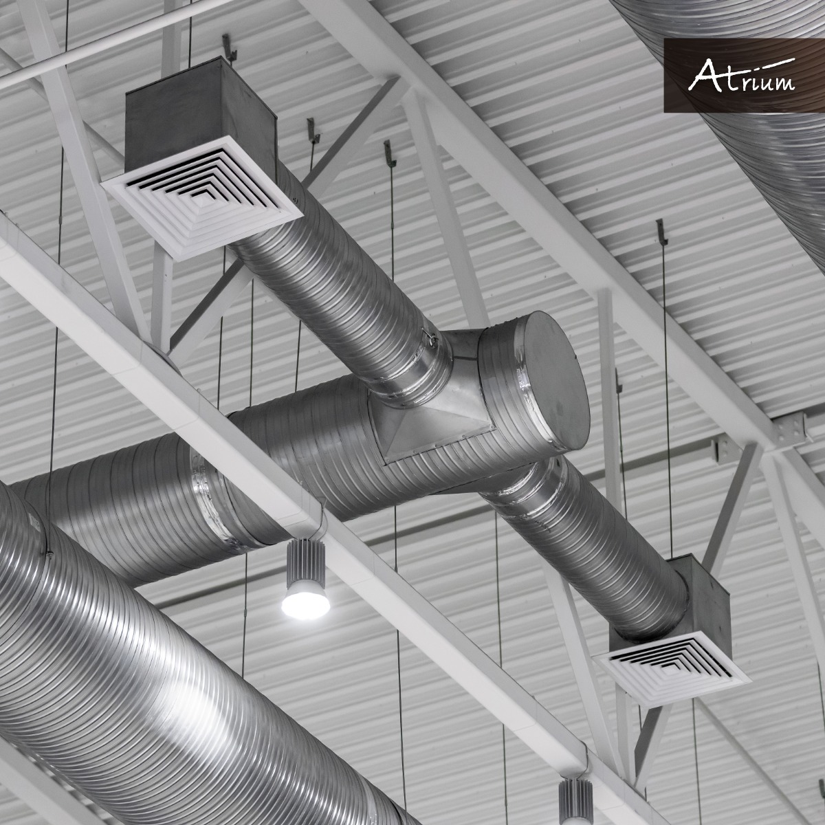 Srebrne, karbowane rury wentylacyjne pod sufitem z białymi belkami konstrukcyjnymi i kratkami wentylacyjnymi, oświetlone punktowymi lampami LED, z logo firmy Atrium w prawym górnym rogu.