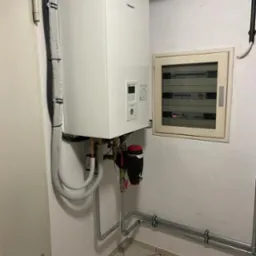 Biały, wiszący kocioł gazowy Panasonic z podłączonymi rurami i czarnym separatorem powietrza, zamontowany na białej ścianie obok skrzynki elektrycznej, z pudełkiem na górze, na tle kafelków...