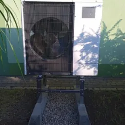 Zewnętrzna jednostka pompy ciepła Mitsubishi Zubadan zamontowana na stalowej konstrukcji z betonowymi podstawami i wysypanym żwirem, na tle zielonej ściany budynku.