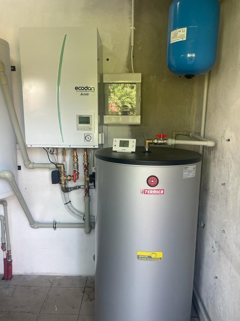 Zainstalowana pompa ciepła Ecodan Mitsubishi Electric z szarym zbiornikiem Termica w pomieszczeniu technicznym z białą ścianą i niebieskim zbiornikiem wyrównawczym.