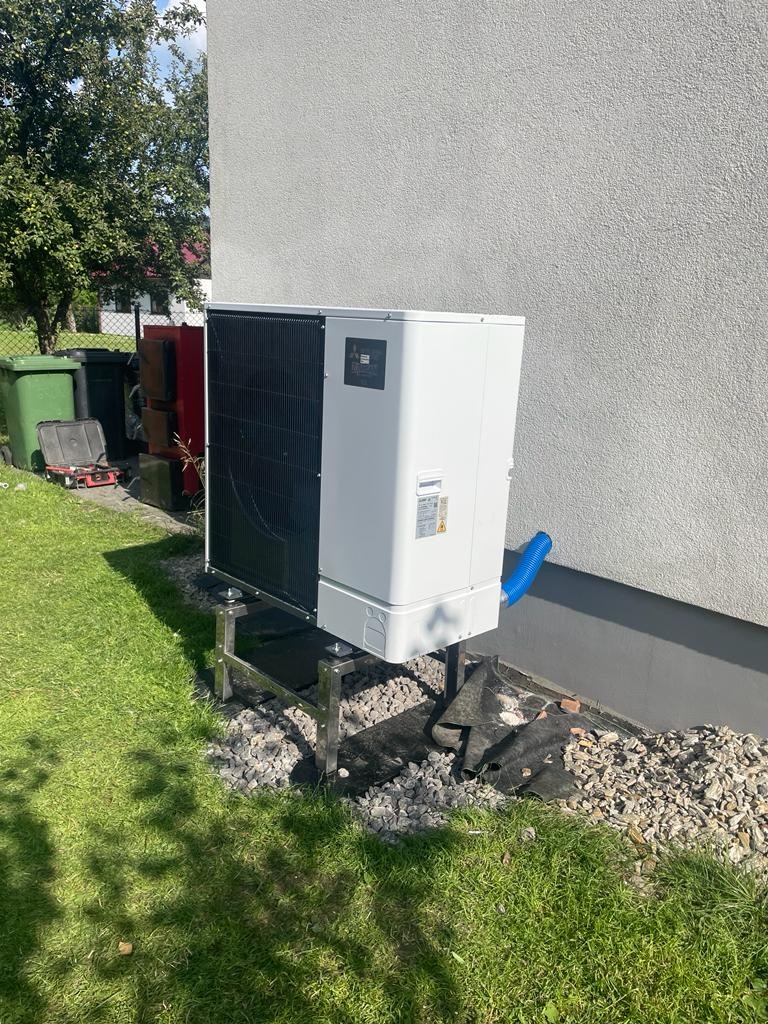 Biała jednostka zewnętrzna pompy ciepła na metalowym stelażu, umieszczona na trawniku przy jasnoszarym budynku, z niebieskim odpływem i widocznymi koszami na śmieci w tle.