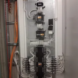 Rozdzielnia sygnału TV/SAT marki Televes z licznymi kablami koncentrycznymi, zamontowana na perforowanej płycie wewnątrz budynku, widoczne także kable w korytkach na ścianie.