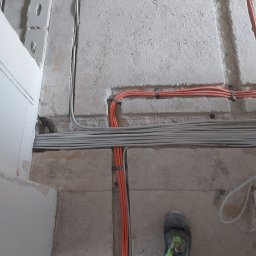 Arkadiusz Bulak - Pomarańczowe i szare przewody ułożone w bruzdach w posadzce z betonu komórkowego, zabezpieczone opaskami, widoczne buty robocze w prawym dolnym rogu.