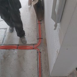 Arkadiusz Bulak - Pomarańczowe przewody elektryczne ułożone w bruździe w podłodze, zabezpieczone czarnymi opaskami, widoczny fragment ściany z zamontowanym domofonem i fragment sylwetki instalatora w roboczych butach.