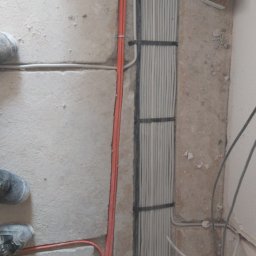 Arkadiusz Bulak - Wiązka szarych przewodów elektrycznych ułożonych pionowo, przymocowanych czarnymi opaskami do ściany, obok czerwone i szare kable w bruzdach w ścianie. Widoczne fragmenty butów roboczych w dolnej...