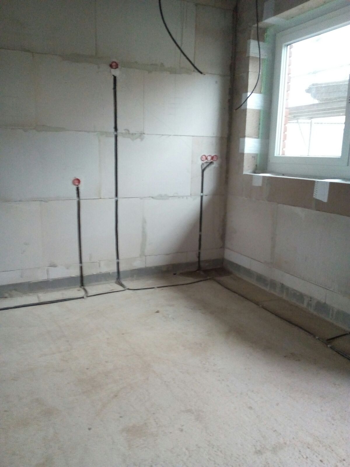Surowy stan wnętrza z widocznymi przewodami elektrycznymi wyprowadzonymi ze ściany, zakończonymi puszkami i gniazdami, instalacja w trakcie realizacji.