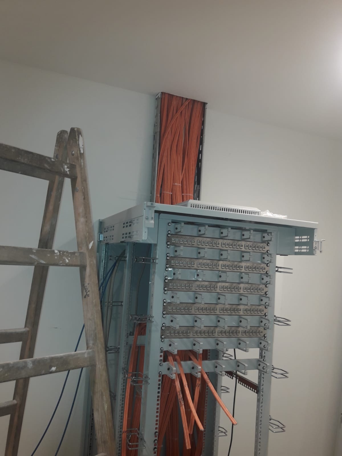 Metalowa szafa krosownicza z licznymi pomarańczowymi kablami, widoczna drabina malarska z boku, kable prowadzone w korycie w ścianie.