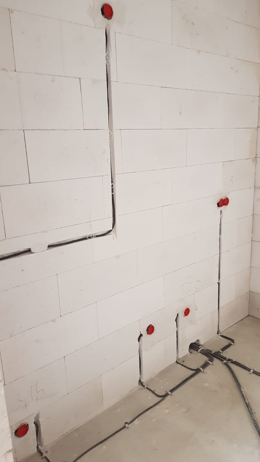 Ściana z betonu komórkowego z wyżłobionymi kanałami pod przewody elektryczne, widoczne czerwone puszki instalacyjne i poprowadzone kable zabezpieczone uchwytami.