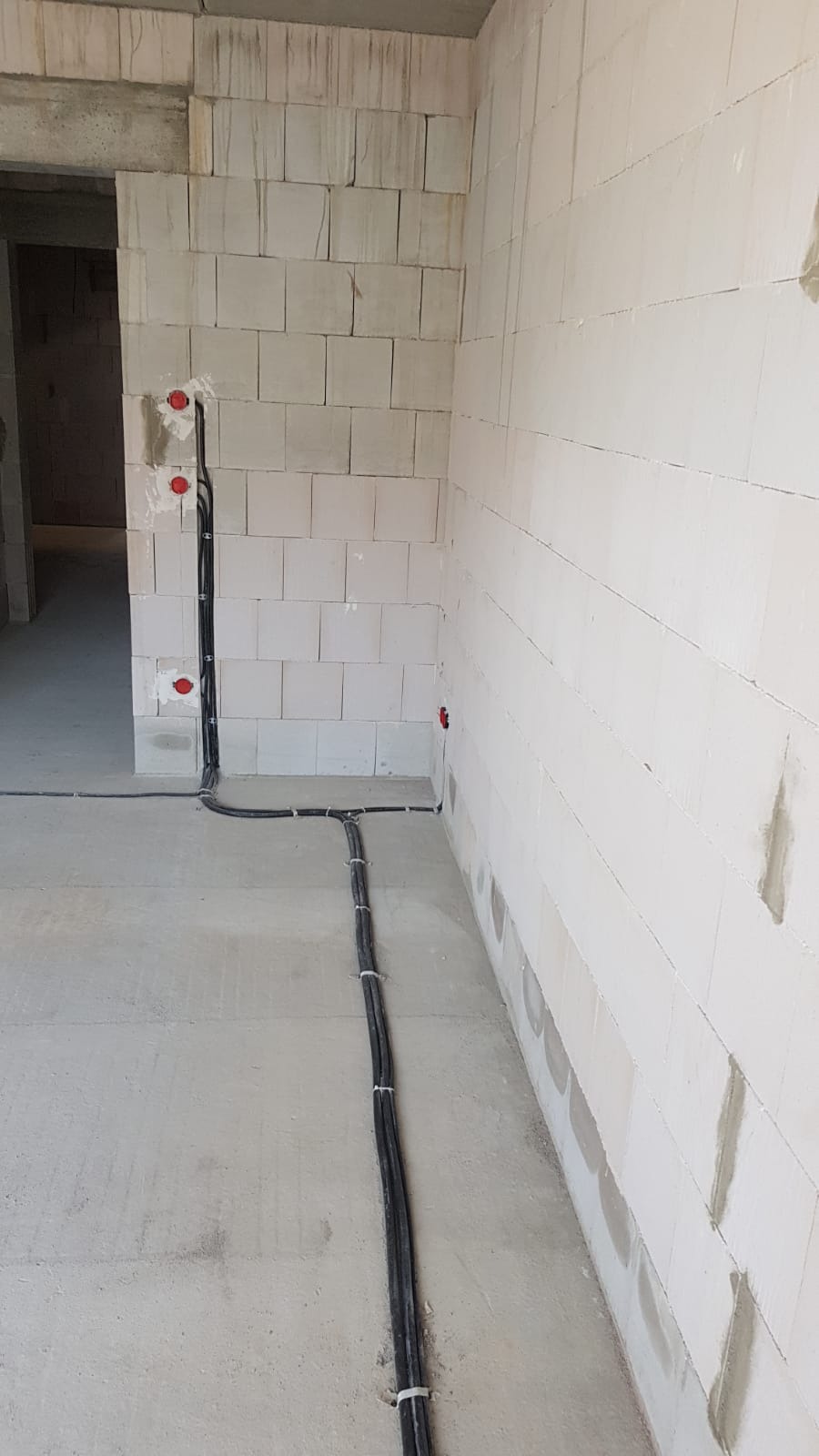 Surowy stan: czarne kable elektryczne prowadzone po podłodze i ścianach z białych bloczków, widoczne czerwone puszki instalacyjne, brak wykończenia.