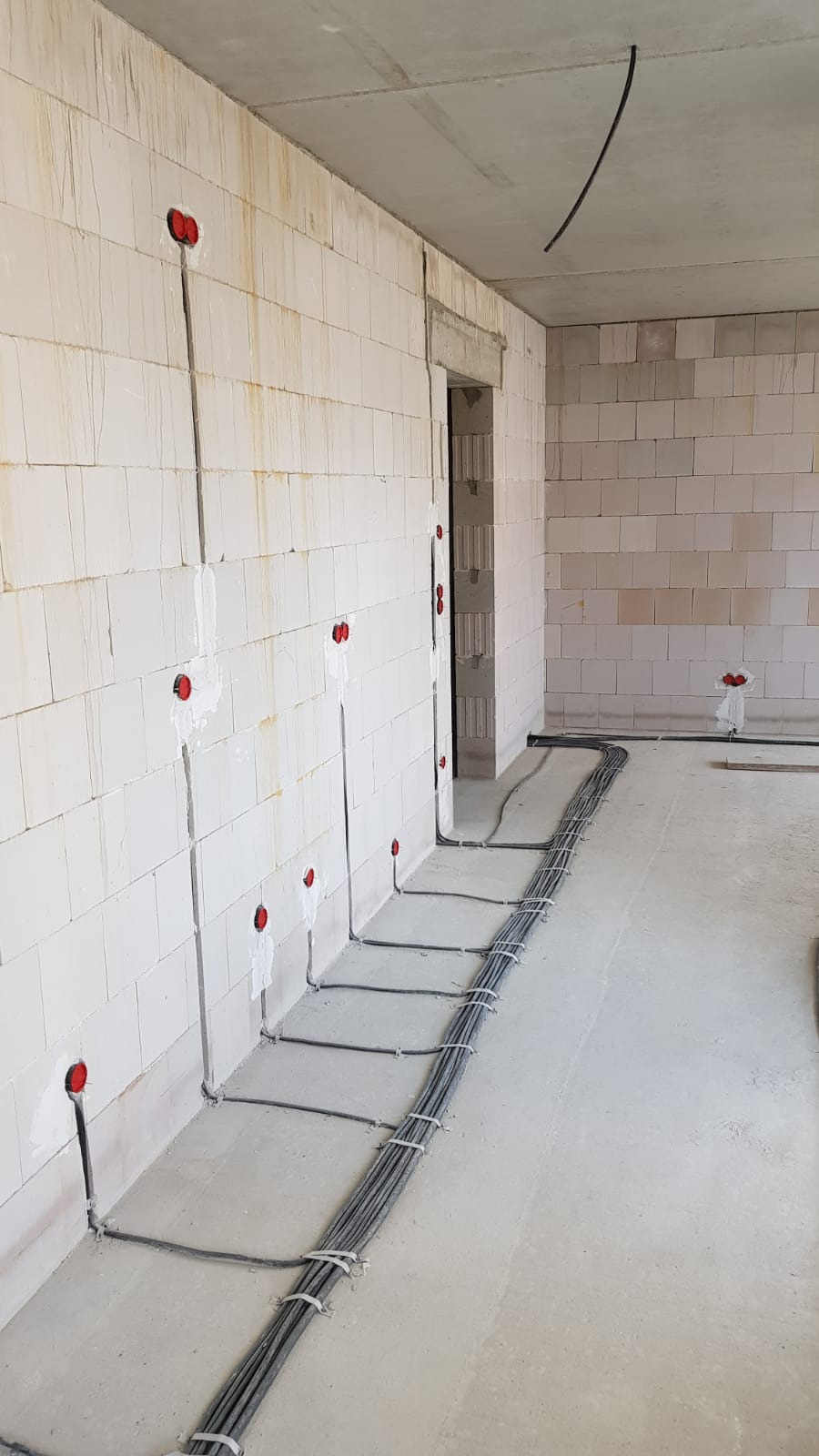 Surowy stan instalacji elektrycznej w nowym budynku: kable prowadzone po ścianach z widocznymi puszkami, wiązka kabli na podłodze tymczasowo spięta opaskami, ściany z białego bloczka.