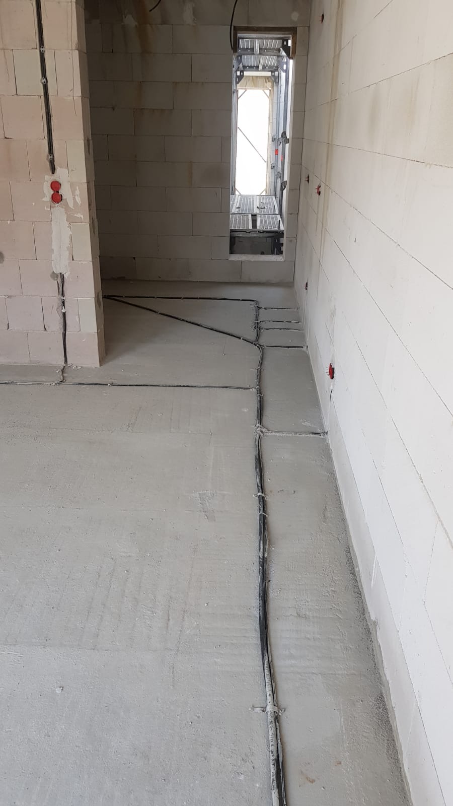 Surowy stan wnętrza budynku w trakcie rozprowadzania przewodów elektrycznych po podłodze i ścianach z widocznymi puszkami instalacyjnymi.