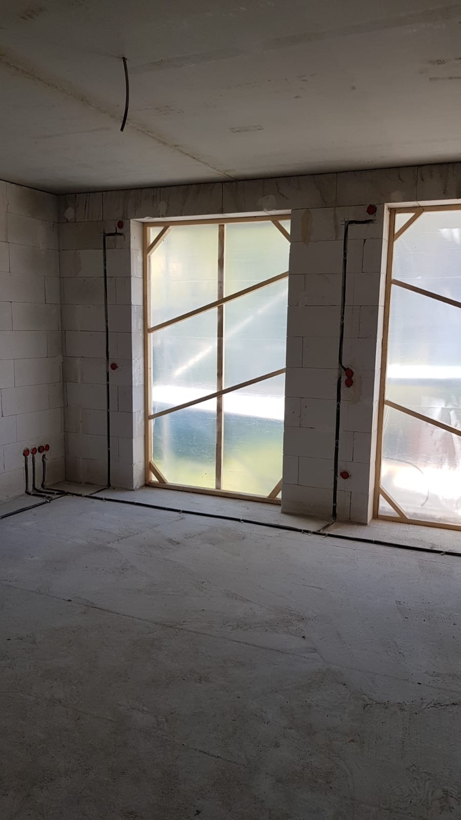 Surowy stan wnętrza z widocznymi przewodami elektrycznymi w ścianach z betonu komórkowego, zabezpieczone okna folią i drewnianymi listwami, oraz wystające ze ściany rurki instalacyjne.