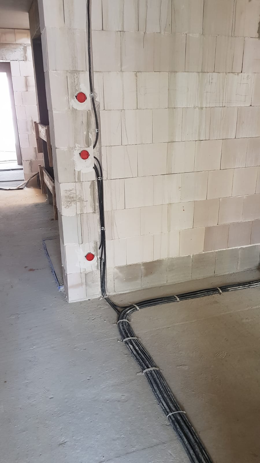 Surowe wnętrze w trakcie budowy: kable elektryczne prowadzone wzdłuż ściany z betonu komórkowego, zabezpieczone obejmami, z widocznymi puszkami instalacyjnymi z czerwonymi zaślepkami.