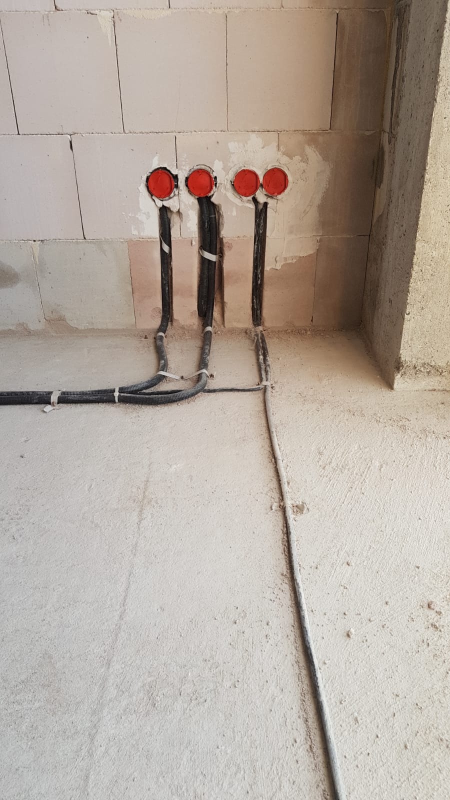 Surowa ściana z betonu komórkowego z trzema przewodami elektrycznymi wychodzącymi z czerwonych puszek instalacyjnych, dodatkowo widoczne przewody w peszlu przymocowane do ściany za pomocą białych...