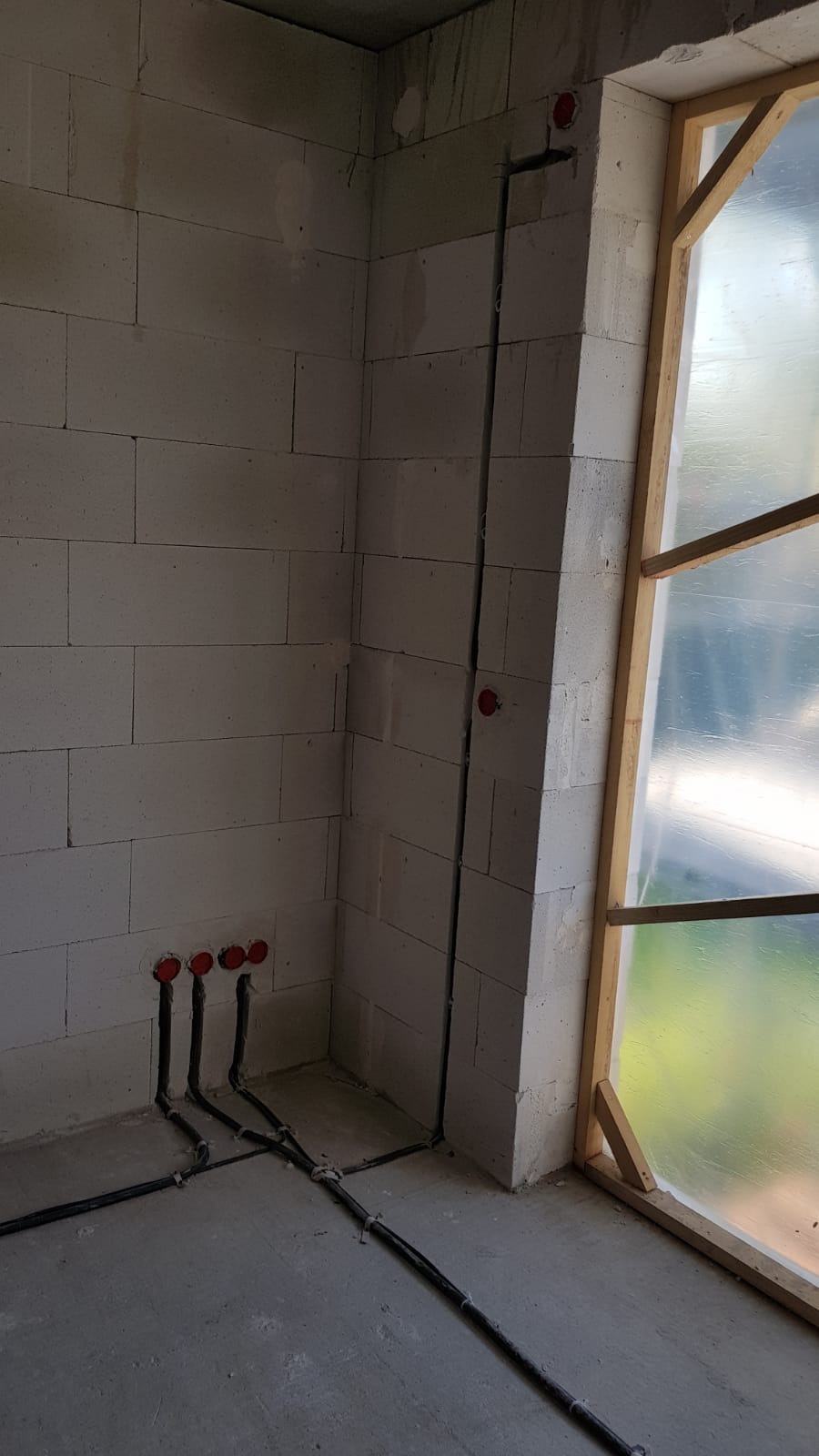 Surowy stan wnętrza z widocznymi przewodami elektrycznymi wychodzącymi ze ściany, zabezpieczone otwory na gniazdka w czerwonym kolorze oraz pionowo poprowadzony przewód wzdłuż ściany obok okna...