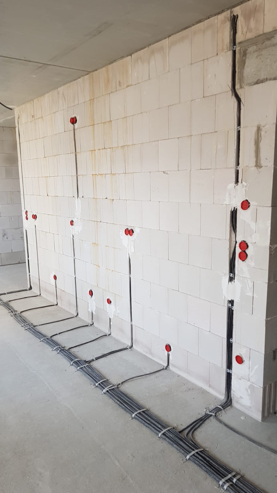 Instalacja elektryczna w surowym stanie: kable prowadzone po ścianie z betonu komórkowego, zabezpieczone uchwytami, widoczne czerwone puszki instalacyjne.