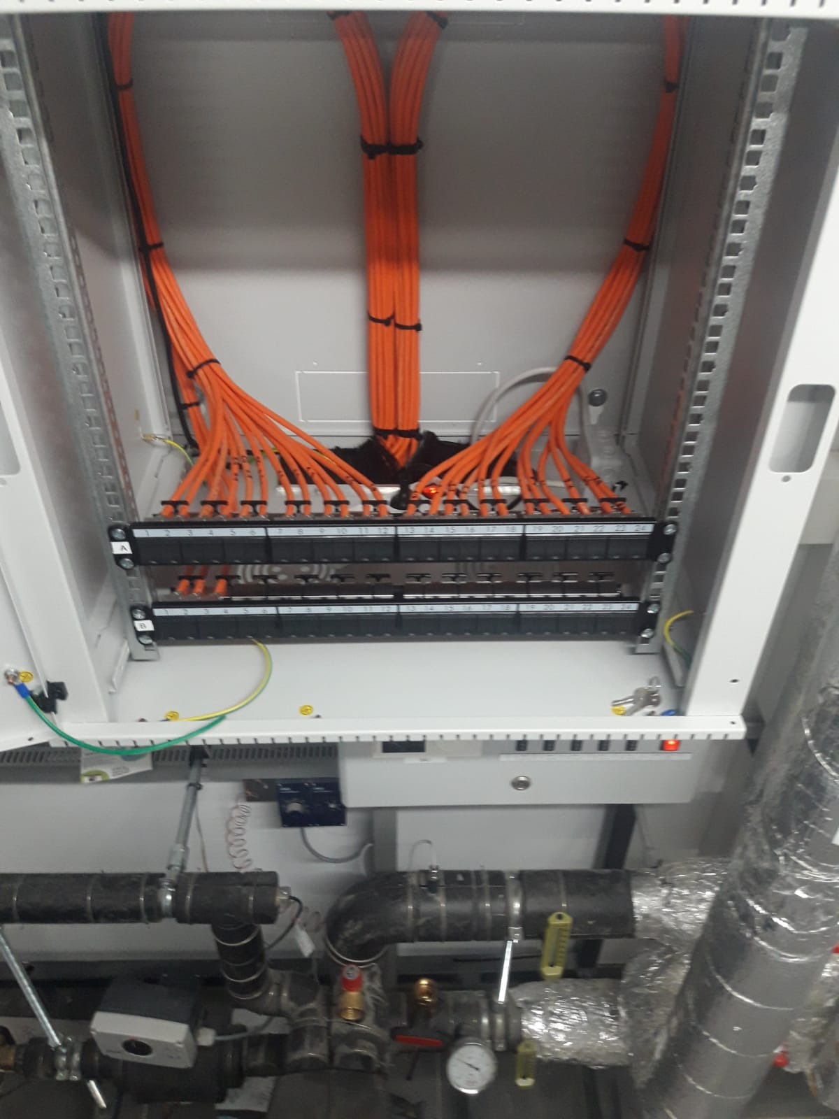 Pomarańczowe kable sieciowe uporządkowane w szafie rack, z widocznymi panelami krosowniczymi i elementami infrastruktury technicznej w tle.