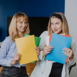 Dwie uśmiechnięte kobiety w biurze trzymają kolorowe teczki, prezentując materiały informacyjne lub dokumenty.