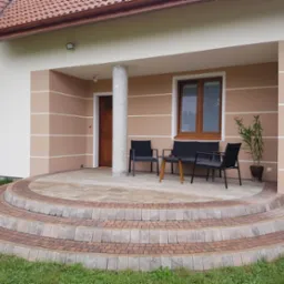 Wykończony taras przed domem w Brzesku z półokrągłymi schodami wyłożonymi kostką brukową w odcieniach beżu i brązu, z meblami ogrodowymi na górze i elewacją w kolorach beżu i bieli.