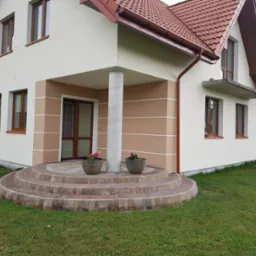 Elewacja domu jednorodzinnego z oknami w drewnianych ramach, schody wejściowe wyłożone kostką brukową, przed kolumną dwa donice z czerwonymi kwiatami.