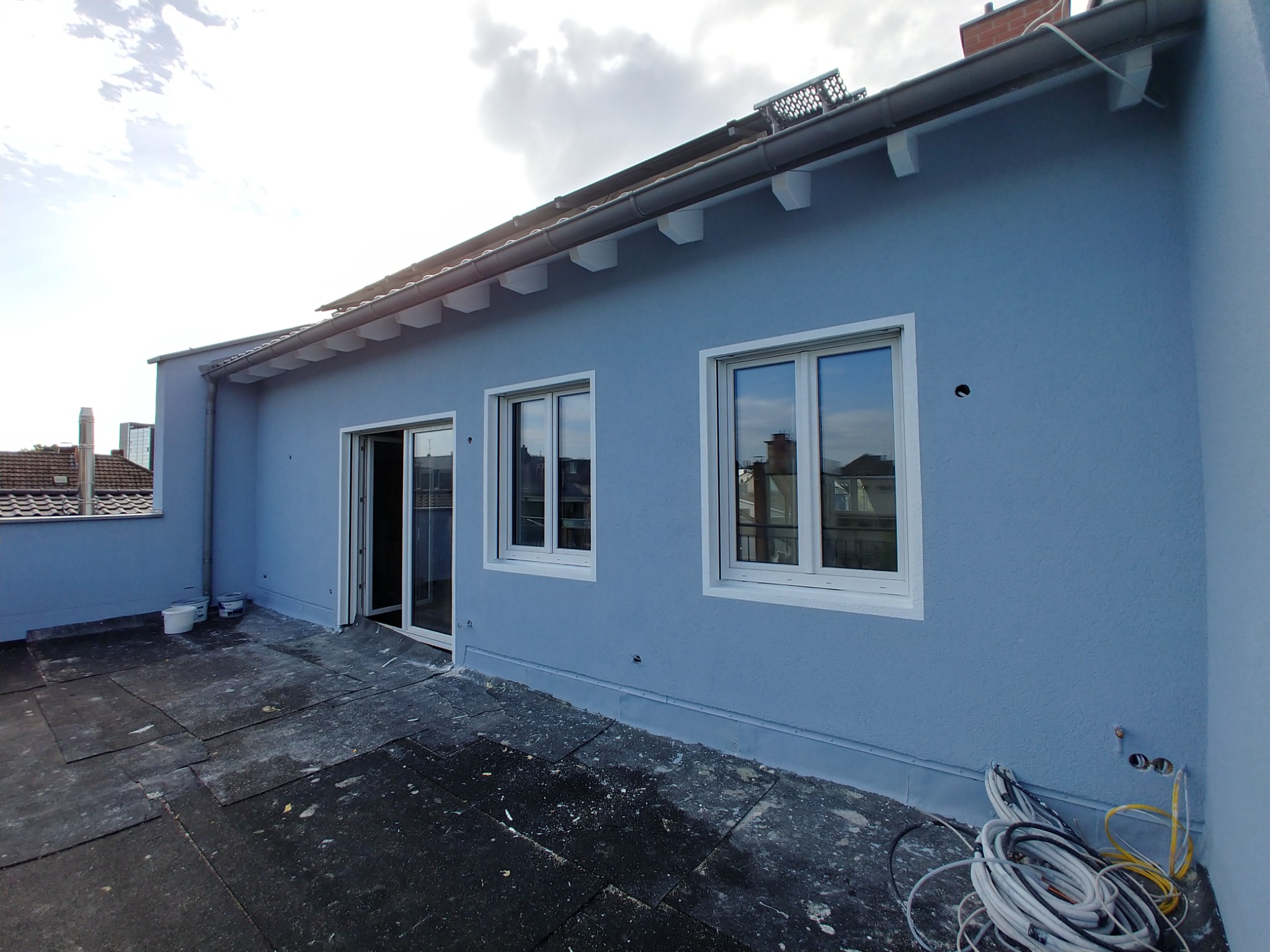 Świeżo pomalowana na błękitno elewacja budynku z białymi ramami okiennymi i drzwiami balkonowymi, widoczne kable elektryczne i puszki instalacyjne na zewnątrz, czarne płyty na dachu, obróbki...