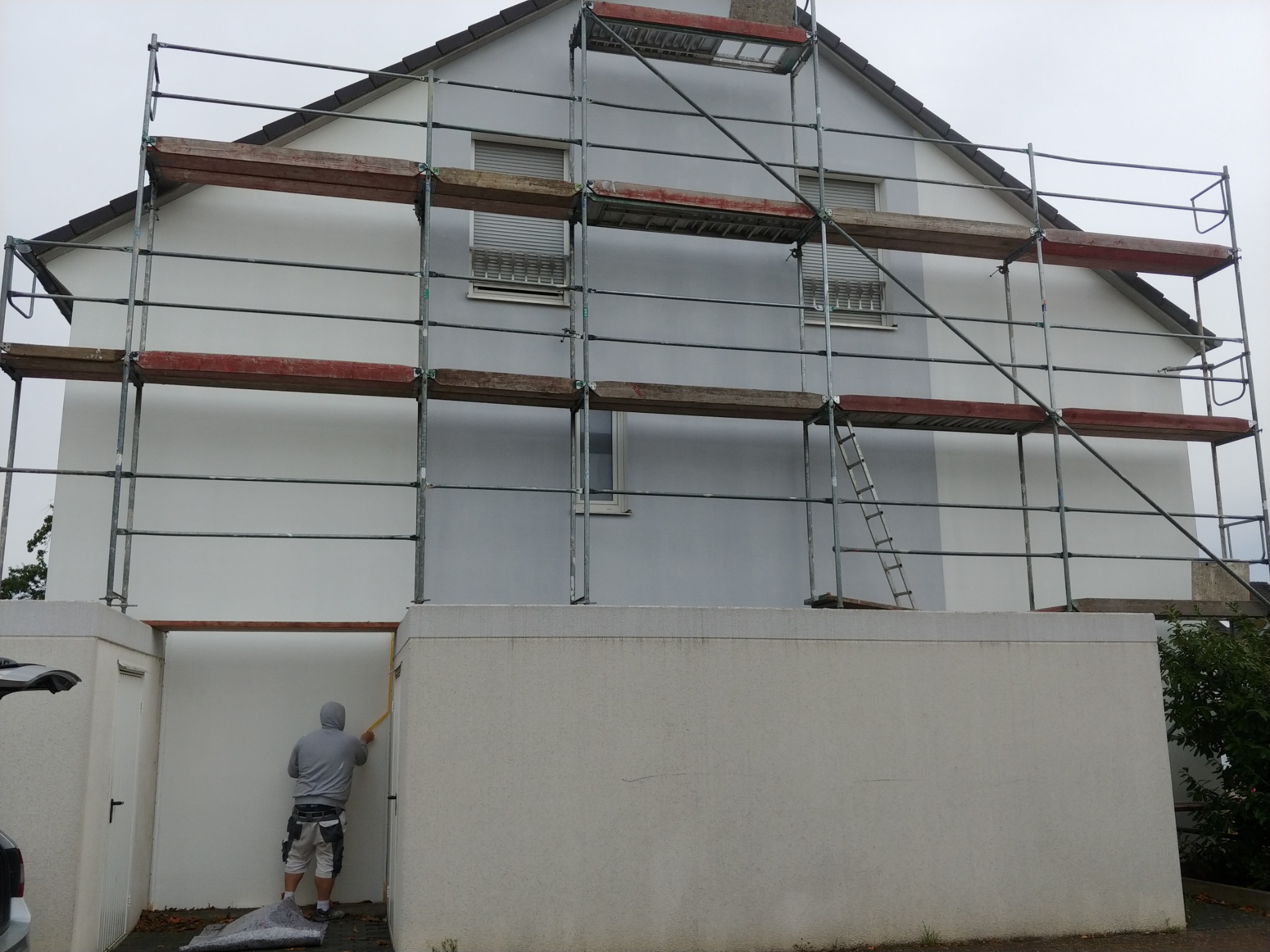 Pracownik w szarej bluzie z kapturem nakłada żółtą taśmę malarską na ścianę zewnętrzną budynku przed malowaniem, widoczne rusztowania i drabina.