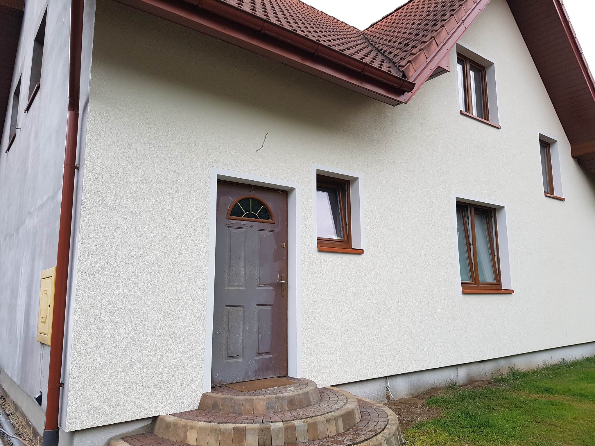 Dom jednorodzinny po ociepleniu elewacji, z widocznymi brązowymi oknami i drzwiami wejściowymi z półokrągłym przeszkleniem, oraz schodami z kostki brukowej prowadzącymi do wejścia.