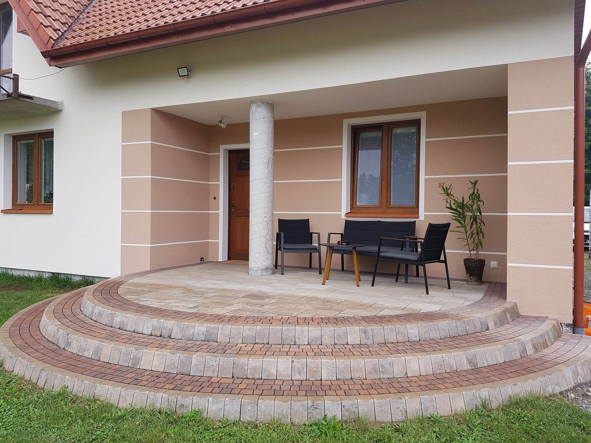 Wykończony taras przed domem w Brzesku z półokrągłymi schodami wyłożonymi kostką brukową w odcieniach beżu i brązu, z meblami ogrodowymi na górze i elewacją w kolorach beżu i bieli.