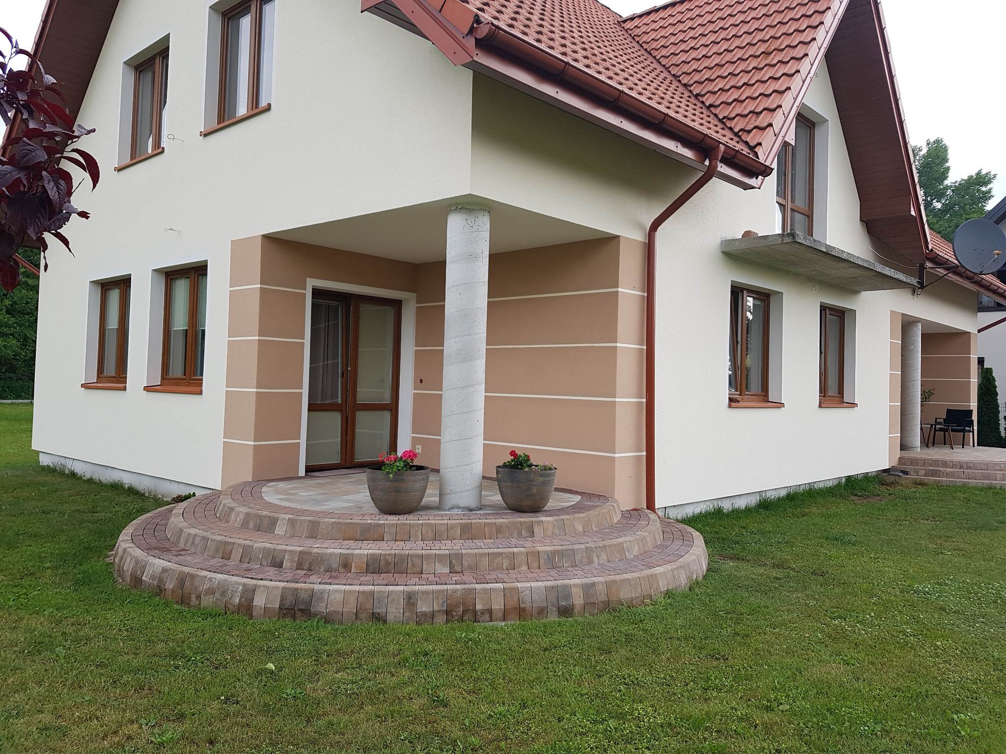Elewacja domu jednorodzinnego z oknami w drewnianych ramach, schody wejściowe wyłożone kostką brukową, przed kolumną dwa donice z czerwonymi kwiatami.