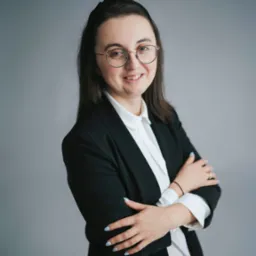 Uśmiechnięta prawniczka w okularach i czarnym żakiecie, z założonymi rękami na tle szarego studia.