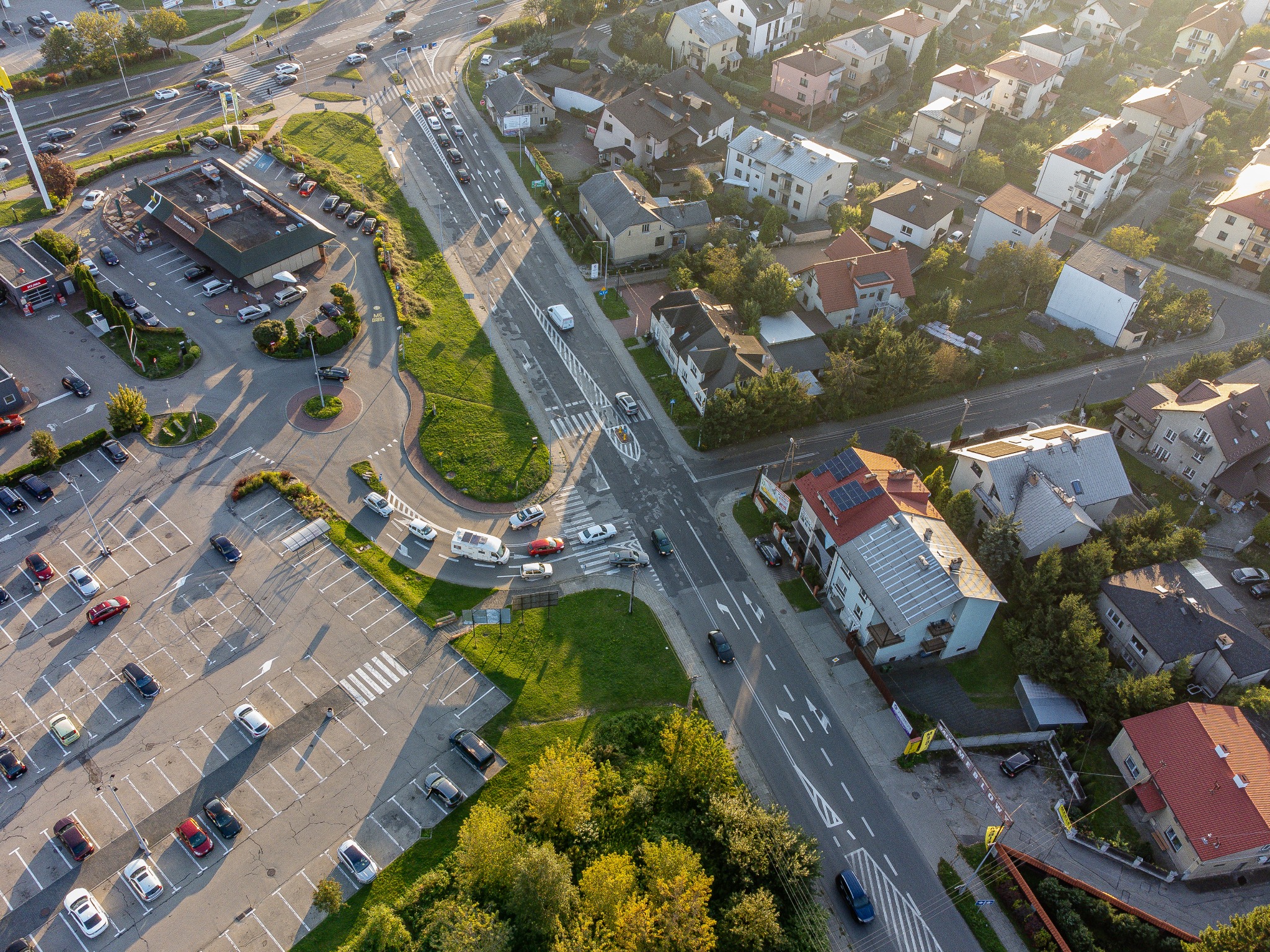 Zdjęcie z lotu ptaka przedstawiające fragment miasta z parkingiem, drogą i zabudową jednorodzinną w słoneczny dzień.