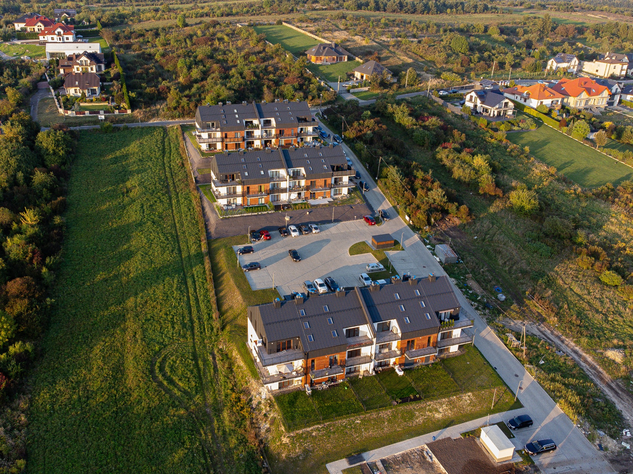 Widok z lotu ptaka na kompleks apartamentowców z parkingiem, otoczonych zielenią i innymi domami w oddali.