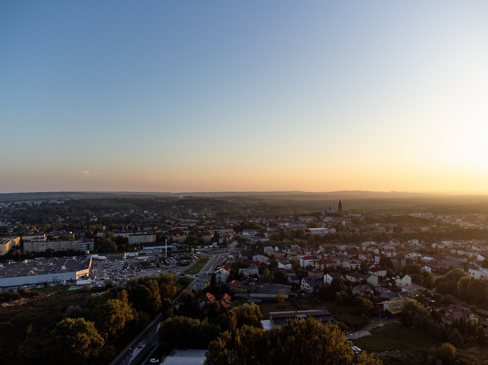Panorama miasta z lotu ptaka o wschodzie słońca, widoczne budynki mieszkalne, sklepy i kościół w oddali.