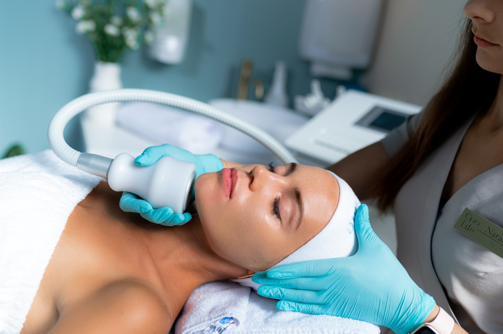 Kobieta poddawana zabiegowi na szyję urządzeniem z głowicą, w gabinecie z białymi kwiatami w tle, wykonywanemu przez osobę w turkusowych rękawiczkach i uniformie.
