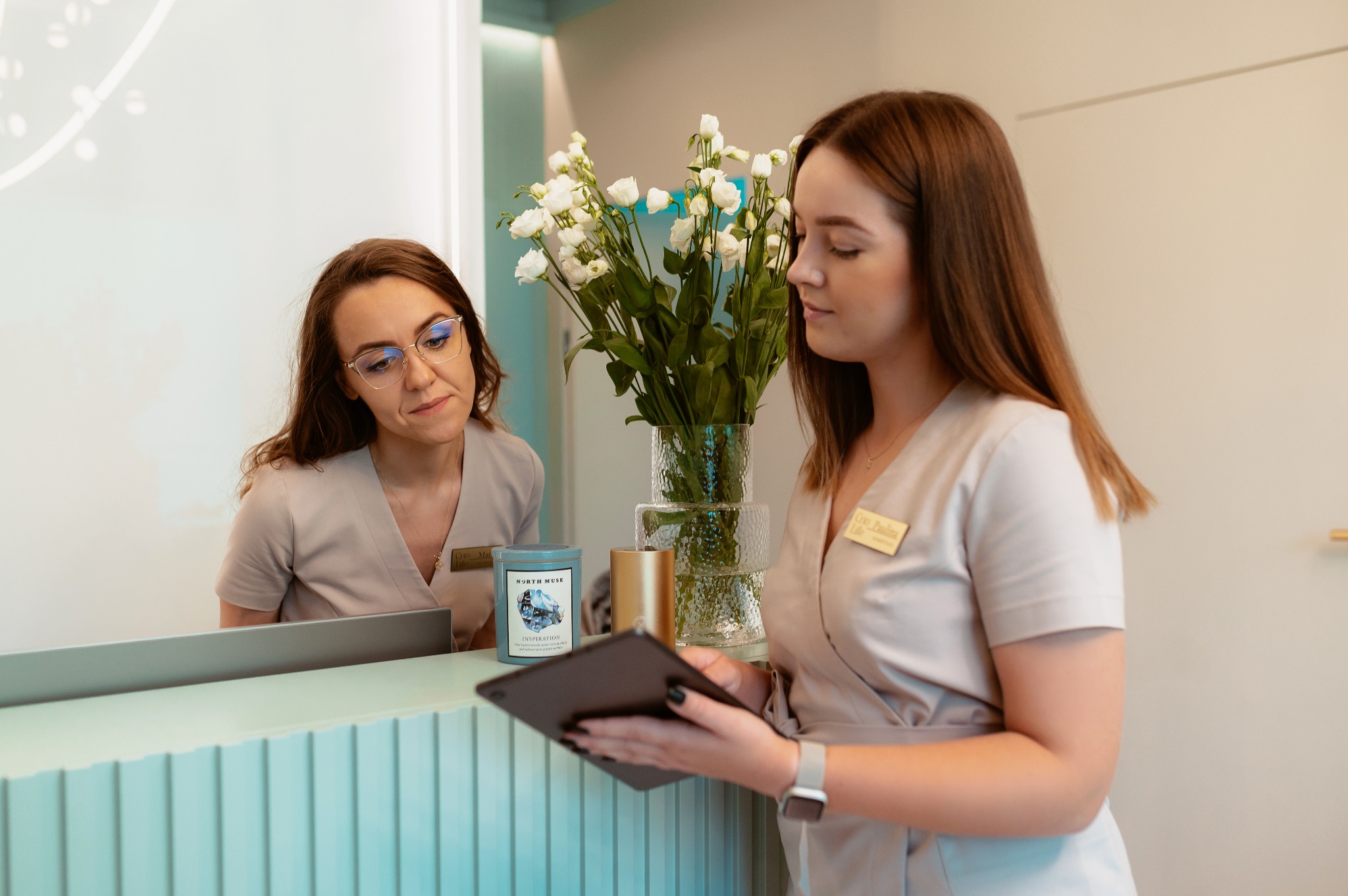 Recepcja gabinetu: dwie kobiety w uniformach, jedna przegląda tablet, obok świeca zapachowa i wazon z białymi kwiatami.