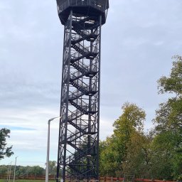 KRY STAL - Wysoka, metalowa wieża widokowa z zewnętrznymi schodami spiralnymi, otoczona drzewami i oświetlona przez nowoczesne latarnie.