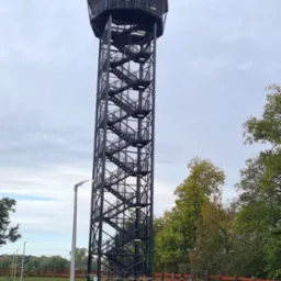 Wysoka, metalowa wieża widokowa z zewnętrznymi schodami spiralnymi, otoczona drzewami i oświetlona przez nowoczesne latarnie.