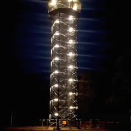 Oświetlona, wysoka wieża widokowa ze stalową konstrukcją i schodami, sfotografowana nocą.