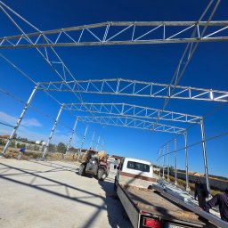 KRY STAL - Stalowa konstrukcja hali w trakcie montażu, widoczny podnośnik teleskopowy i samochód dostawczy z elementami konstrukcyjnymi.