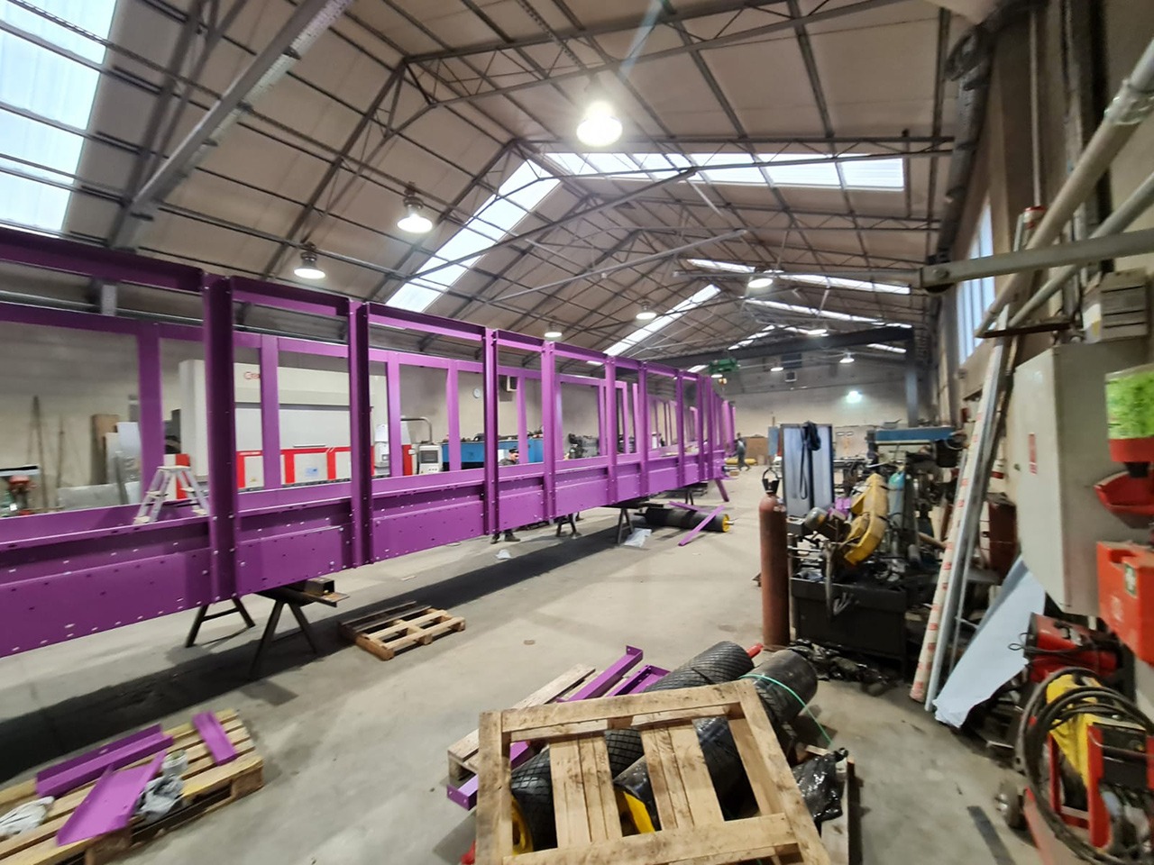 Fioletowa, stalowa konstrukcja ramy w hali produkcyjnej, wsparta na kozłach, widoczne palety i sprzęt warsztatowy w tle.