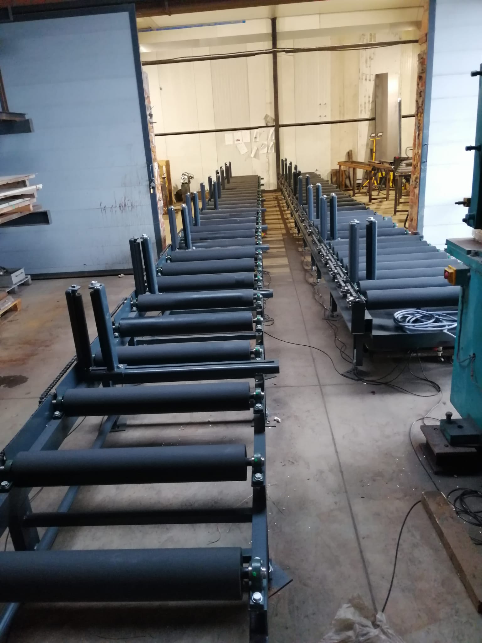 Długi, metalowy system transportu rolkowego z czarnymi rolkami w hali produkcyjnej, z widocznymi elementami konstrukcyjnymi i przewodami.