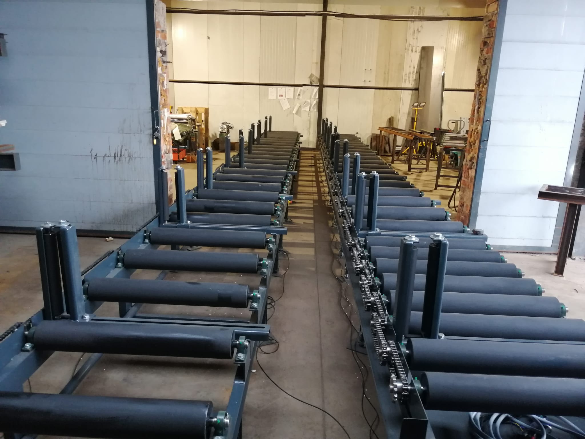 Długi, dwutorowy system transportowy z czarnymi rolkami i łańcuchami napędowymi, ustawiony w hali produkcyjnej z widocznymi elementami konstrukcyjnymi i oświetleniem przemysłowym.